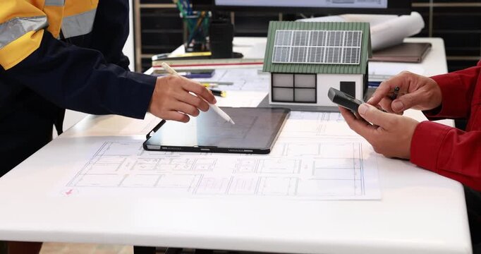 Engineers review house plans with a solar panel model, tablet and smartphone on a desk, discussing sustainable home design and renewable energy.
