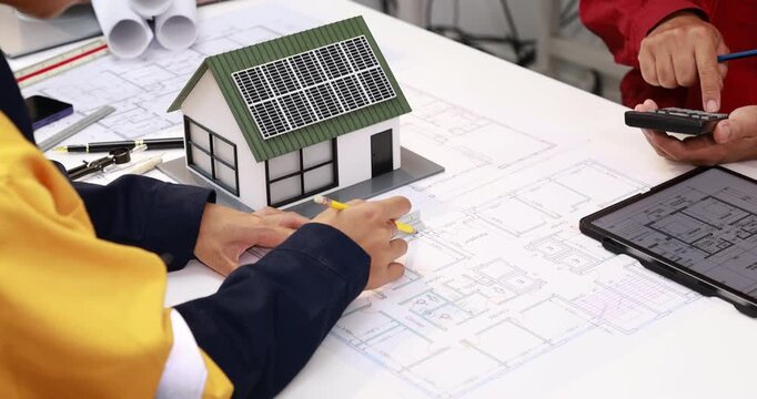 Architects review house plans beside a model home with rooftop solar panels, discussing sustainable building design at a desk.