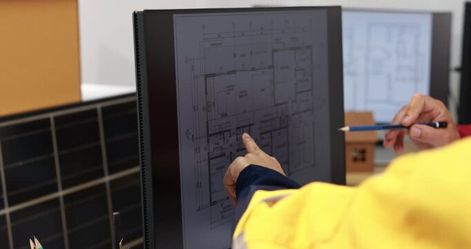 Engineer in a yellow safety jacket points at building plans on a computer monitor while holding a pen in a modern office.