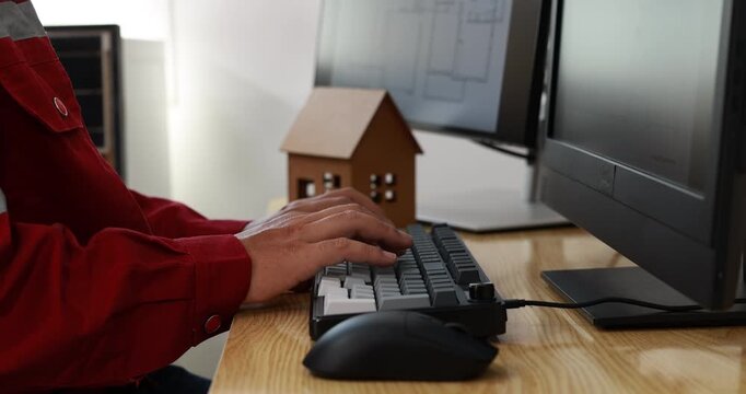 Close view of a person in a red shirt typing on a keyboard at a wooden desk with dual monitors, mouse, and small house model, suggesting real estate work.