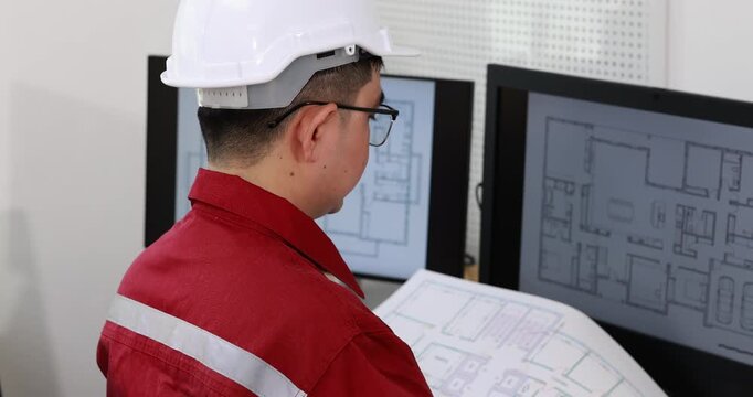 Rear view of an engineer in a hard hat and red work uniform reviewing building plans beside computer screens with floor layouts.