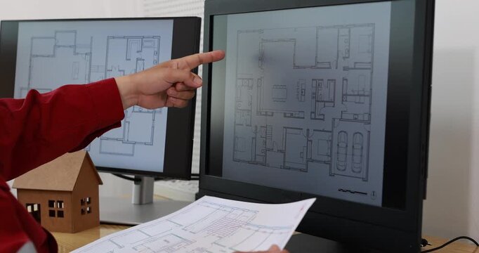 Architect pointing at house floor plans on computer monitors beside a small model home, reviewing building drawings in an office.