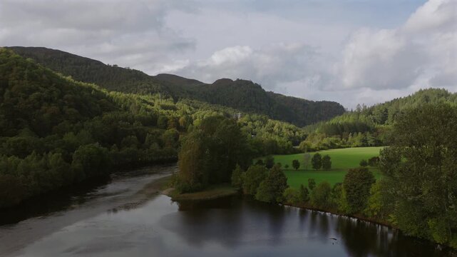 Drone shot of the River Garry showing the river and woodland in the background. As the shot rises a small hydro electric generator is seen next to the river along with pylons.