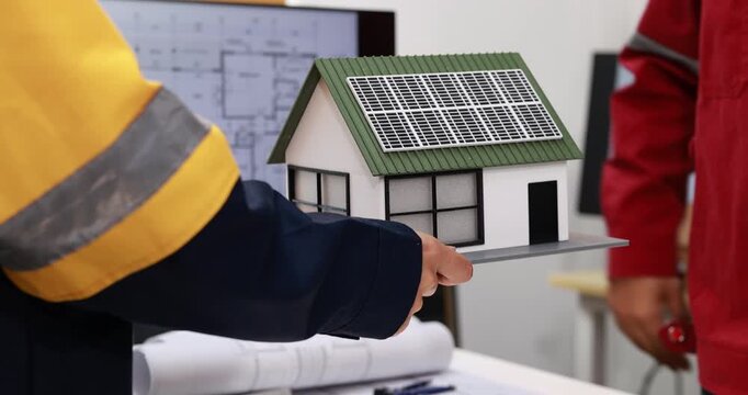 Construction workers review a model house with rooftop solar panels and building plans, renewable energy planning and home project design.