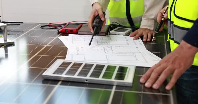 Engineers in safety vests review building plans and solar panel samples on a table, planning a renewable energy installation.