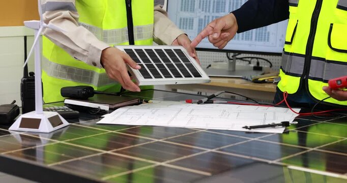 Engineers in safety vests inspect a portable solar panel and electrical equipment on a desk with solar modules, plans and computer screens.