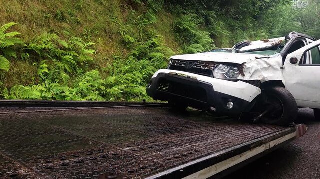 Roadside assistance loading a completely destroyed white car onto a flatbed tow truck after a serious traffic accident on a country road