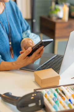 Mature Asian woman holding smartphone and typing on laptop manage online store handcrafted jewelry