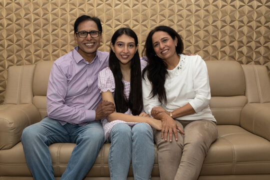 Indian happy mature couple and young girl are hugging, looking at camera and smiling while sitting on couch at home. Concept of Happy Daughter Day.