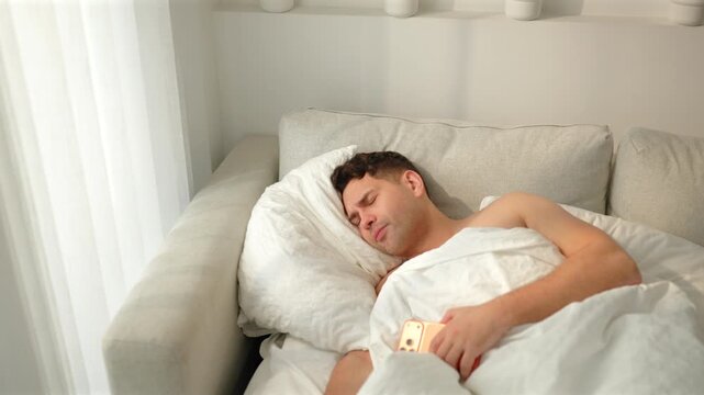 A groggy shirtless man rubs his forehead on a white pillow, then sits up frowning and squinting at an orange phone. Lazy morning wake up on a gray sofa bed by sheer white curtains in bright sunlight.