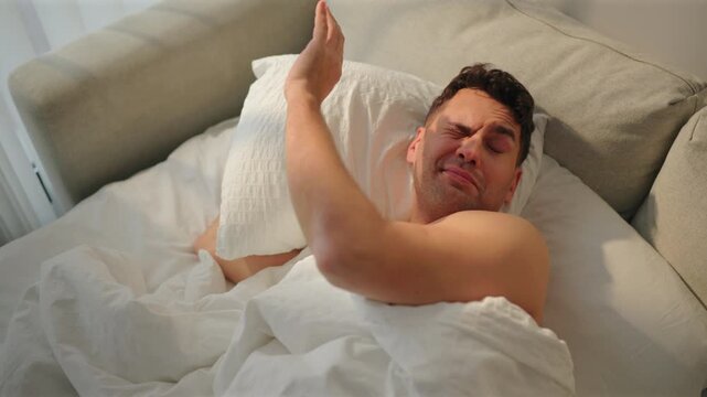 Shirtless man shields his face from bright morning sun with a raised hand, then yawns with mouth wide open and stretches both arms. Tired wake up on gray sofa bed under white duvet by sheer curtain.