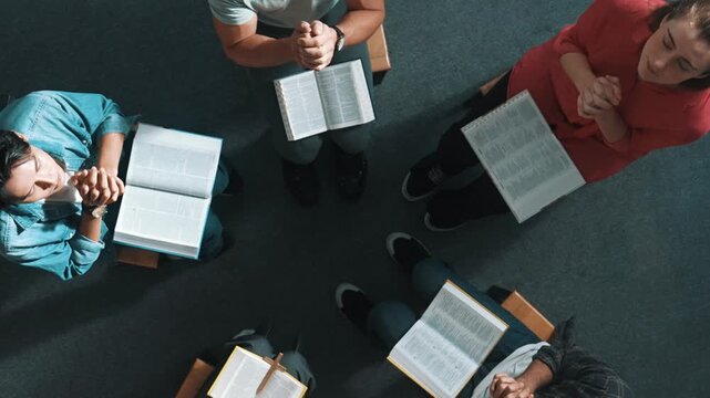 Top down view of prayers reading bible with cross on the laps while making folded hands. Aerial view of diverse people praying to god with faith, trust and hope. and sitting in circle. Symposium.