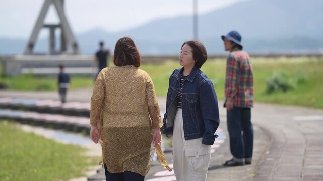 Soft light bathes a tranquil park path. Women walk, chatting, heads tilted upward. Man strolls behind, gaze fixed ahead. Mountains loom gently in the distance. Ideal for serene outdoor, travel