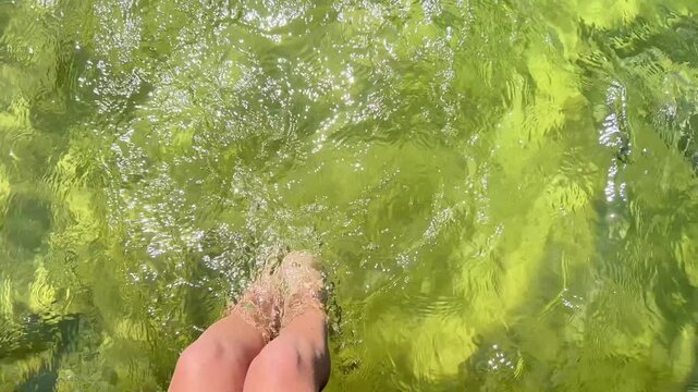High-angle shot of a person dangling legs and bare feet in clear, sunlit shallow water. Ideal for travel promos, summer vacation ads, wellness, and leisure lifestyle videos.