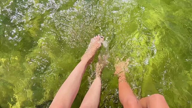 Top-down shot of two people dangling bare feet and splashing in clear, sunlit river water. Ideal for summer vacation, travel, eco-tourism, and leisure lifestyle videos.