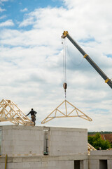 Seen from below, a construction crane is lifting a wooden roof truss above unfinished masonry walls at a building site beneath cloudy skies. © vladim_ka