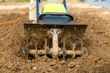 Rotating tiller is aerating and mixing rich soil in a garden bed for effective planting. The machine is actively working to prepare the earth, showing its rotating blades, outdoors. © vladim_ka