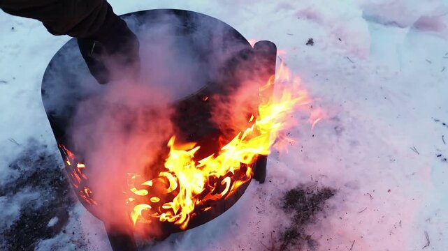 Close-up of outdoor cooking in winter, with food simmering in pan above decorative metal stove and bright fire glowing through the patterned sides. Traditional rustic food preparation in cold weather.