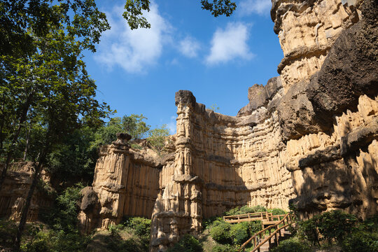 Slender eroded pillar fronts curving layered cliffs with wooden steps and trees at Pha Chor, Mae Wang National Park, Doi Lo, Chiang Mai, Thailand. Upward view shows sediment grooves and blue sky.