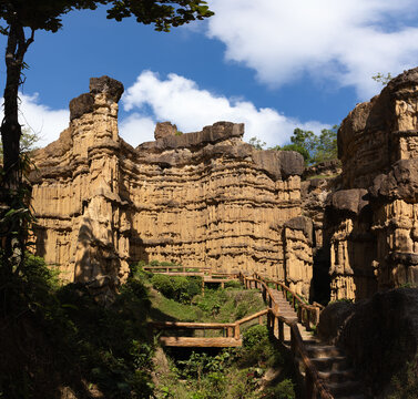 Jagged sediment towers and banded cliff walls surround wooden stairs under white clouds at Pha Chor, Mae Wang National Park, Doi Lo, Chiang Mai, Thailand. Leafy canopy frames sunlit geologic scenery.