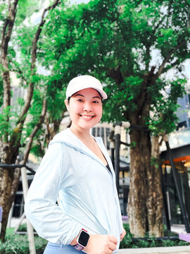 Portrait of a smiling Asian woman in light blue activewear and a white cap standing outdoors in a modern urban park