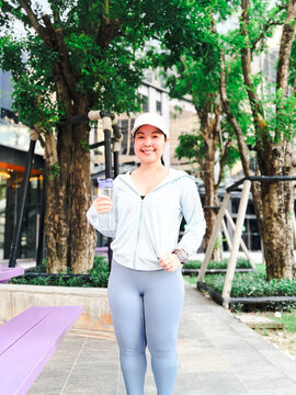 Portrait of a smiling Asian woman in light blue activewear and a white cap holding a water bottle while standing outdoors in a modern urban park