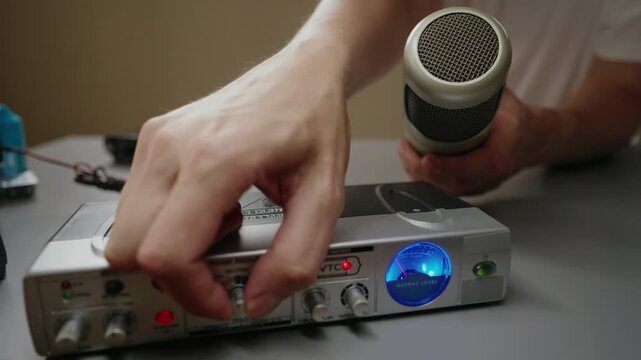 Close-up of male hand adjusting knob on professional audio preamplifier, holding condenser microphone, preparing for recording session in studio, output level meter lit, close-up, slow motion.