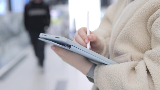Person holds tablet, uses stylus in bright indoor space. Background shows blurred people walking past. Focus remains on hands and device throughout. Outfit appears cozy, light-colored