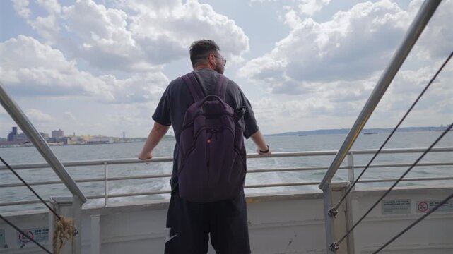 Man with purple backpack leans on ferry stern rail in New York City, New York, USA. Back view of traveler watching white wake, Brooklyn shore and Verrazzano Bridge under summer clouds on a sunny day.