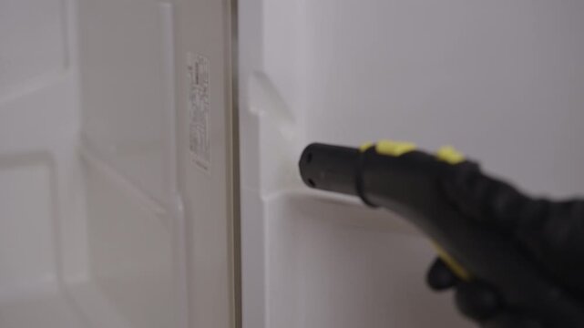 Housewife wearing black protective gloves using powerful steam cleaner to sanitize inside of refrigerator, wiping surface with paper towel for perfect result. Concept of cleanliness and housekeeping.