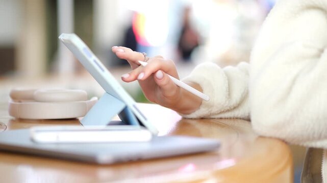 Person uses tablet with stylus on wooden table. Warm lighting highlights hands and device. Background softly blurred with indoor setting. Table holds notebook and coffee cup