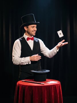 Magician performing a card trick on a theater stage. Playing card floats above a top hat and red-covered table. Magic show entertainment. Illusion, sleight of hand and live performance.
