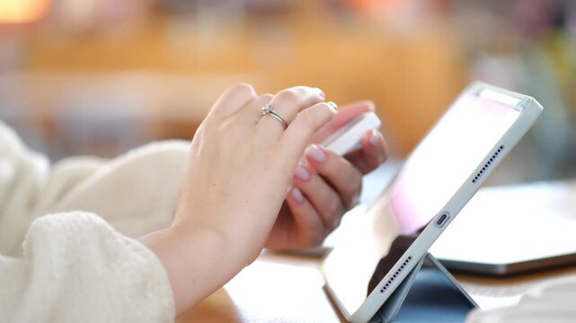 Person uses tablet in cozy indoor setting. Hands type on screen with focus. Background blurred with warm tones. Casual clothing visible. Modern tech in daily use