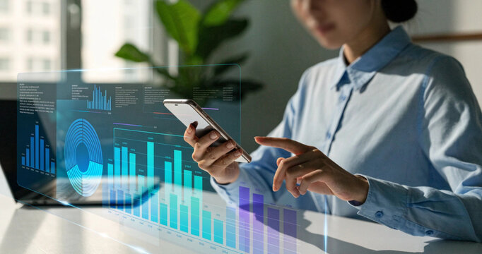 Businesswoman using smartphone with virtual financial dashboard hologram in bright modern office. Digital financial analytics screen displaying blue bar charts and data statistics for business growth