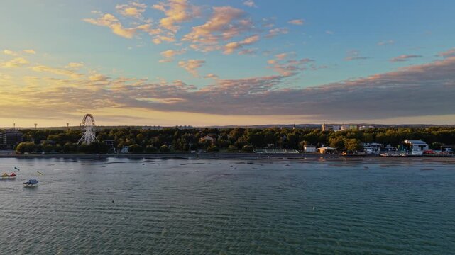 Lake Balaton glimmers at sunset while long rowing boat moves across water. This broad lakeside view includes tree lined shore, distant buildings and Ferris wheel beneath colorful clouds.