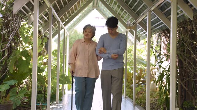 Senior woman uses walking cane beside young man offering steady support along garden walkway while encouraging independence, safe mobility through care, physical support, companionship.