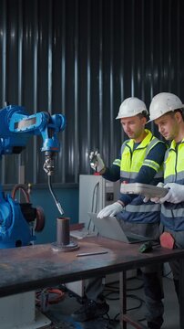 Two workers inspect machinery beside a blue robotic arm. They wear safety vests, gloves, and hard hats