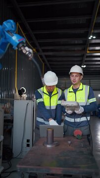 Two workers inspect machinery beside a blue robotic arm. They wear safety vests, gloves, and hard hats