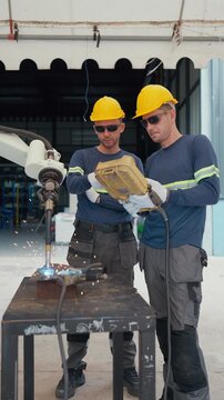 Two welders in hard hats work beside a metal table. One man holds a welding torch, sparks fly from the cut. Both wear gloves, sunglasses