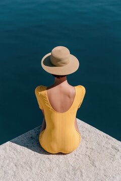 Overhead view of a woman in a yellow striped swimsuit and straw hat sitting on a stone ledge above deep blue water for summer wallpaper and background