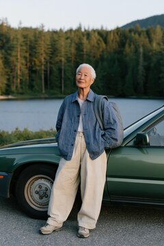 Senior person leaning on green car by a forest lake in casual cuffed trousers for lifestyle poster and outdoor background