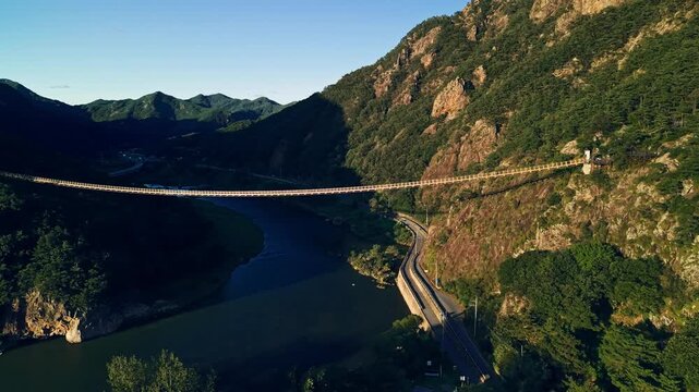 Aerial view of Wolyeongsan suspension bridge spanning Geumgang River between rocky mountains and winding cliffside road in Geumsan Korea at golden hour