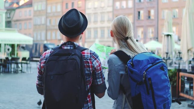 Three tourists with backpacks stand in a European city square during daytime, examining a map while surrounded by outdoor cafes and traditional architecture.