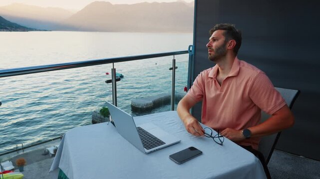 Thoughtful remote worker relaxing on balcony with laptop and smartphone, enjoying peaceful sunset view over calm lake and mountains after a long workday