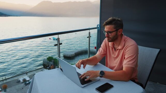 Focused remote worker using laptop on peaceful seaside balcony at sunset, balancing productivity and relaxation with ocean view and modern outdoor workspace