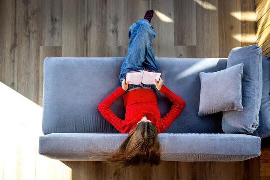 Young woman in red sweater lying upside down on gray sofa while reading an open book, overhead view of relaxing leisure time in a cozy modern home with natural sunlight.