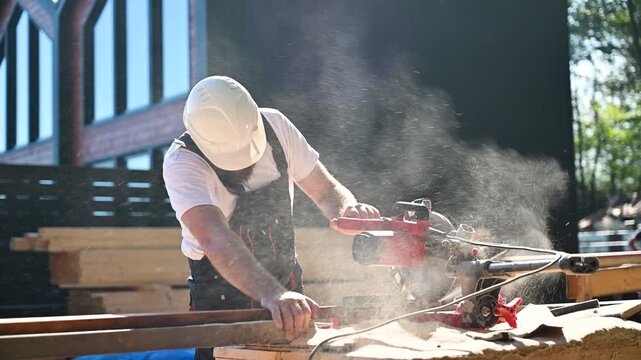 Worker saws lumber on power miter saw outside residential building. Close action shot of wood cutting, flying dust, and professional construction process