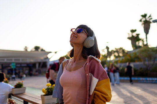 A woman wearing headphones and sunglasses enjoys her favorite song on a sunlit street. The woman smiles as she listens to the music and shows off her rings and bracelets.
