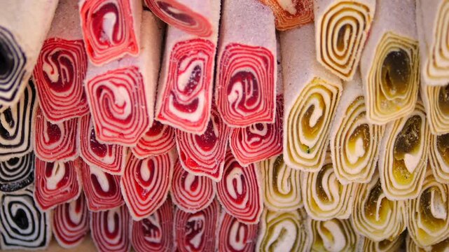Colorful rolled candy sticks hang in dense rows, revealing bright spiral cross sections. Striped sweets in red, black and gold create vivid assortment of decorative confectionery arranged in close
