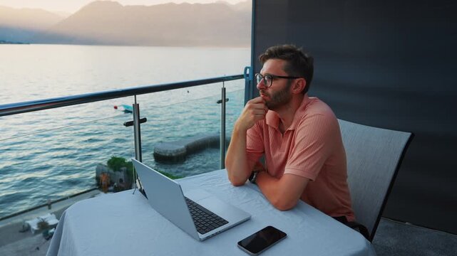 Man working remotely on balcony with laptop overlooking tranquil waterfront at sunset, reflecting thoughtfully during online communication or focused freelance work session
